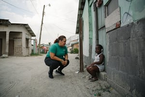 Woman in a OneChild t-shirt stops to talk with a toddler sitting by a concrete building