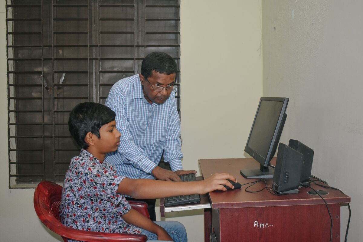 A boy seated at a computer receives guidance from a teacher standing next to him. 
