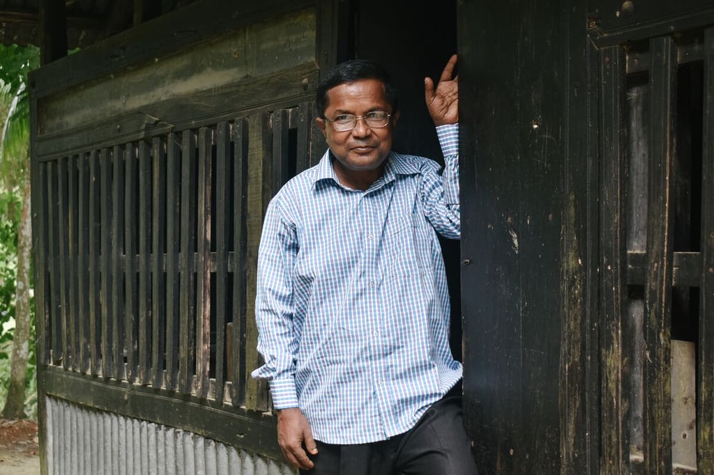 A man in glasses and a blue shirt stands in the doorway of a rough wood building in Bangladesh. 