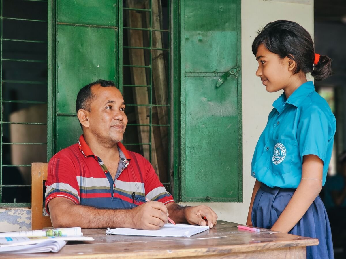 A teacher in a red shirt grades papers at a table and talks with a girl in a blue school uniform. 