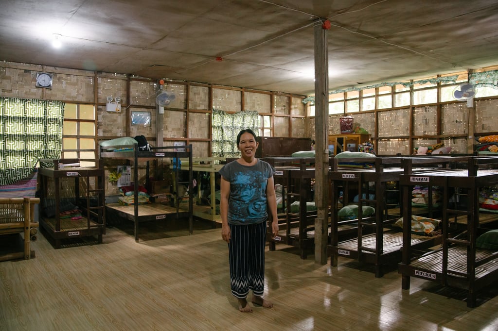A smiling Filipino woman stands in a doorm room with woven mat walls and many bunk beds