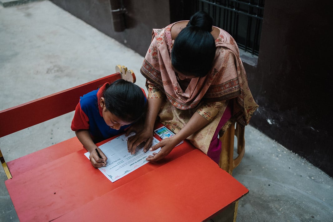 A woman in pink and gold Indian clothing helps a girl writing at ta desk.