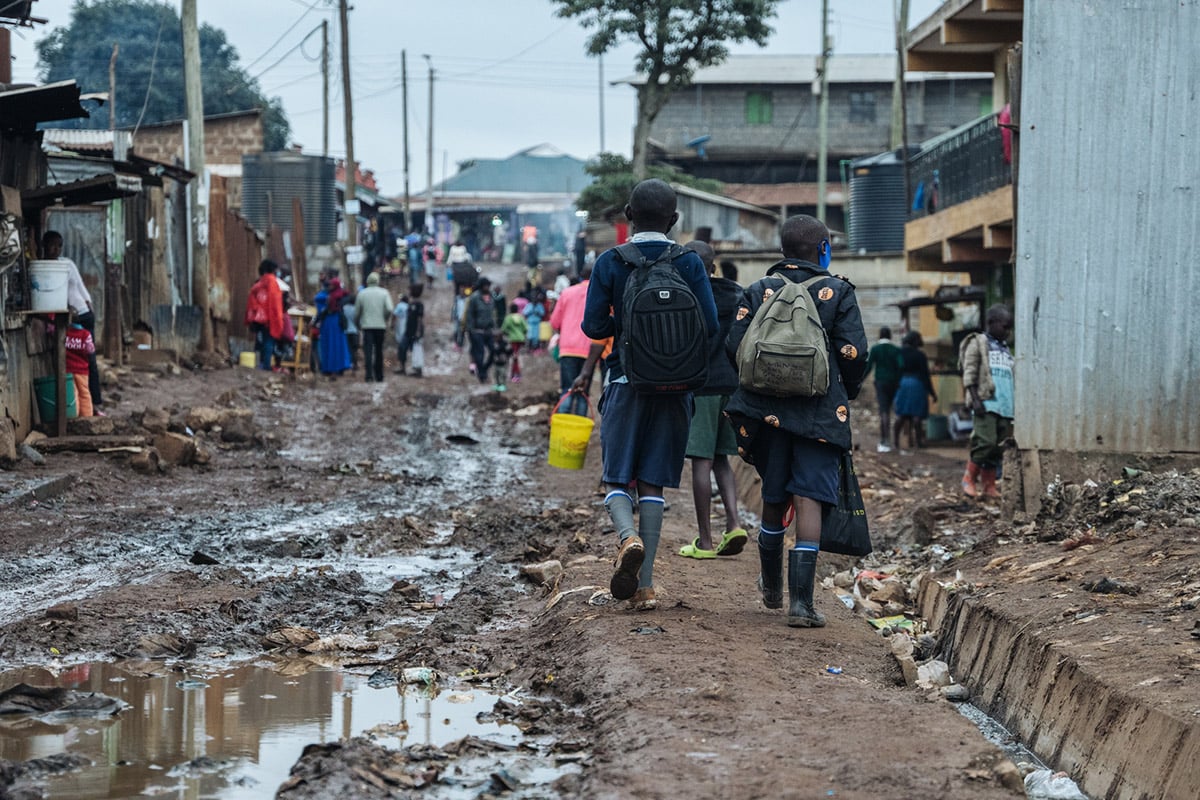 Two children with backpacks walk along a muddy path through a slum in Kenya